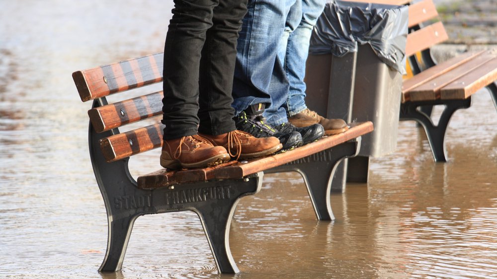 Three people standing on a bench in an area that has flooded. Brown flood water threatens their safety.